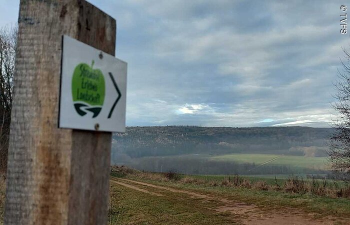 Blick auf einen grünen Berg in der herbstlicher Landschaft. Im linken Bildrand ein Holzposten mit einem Schild. Ein grüner Apfel auf weißem Grund mit der Aufschrift Streuobst Erlebnislandschaft.