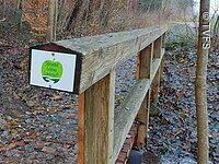 Eine Holzbrücke bedeckt mit Herbstlaub führt entlang von Bäumen und Sträuchern. Der Boden ist nass vom Regen. Am Brückengeländer Hämgt einSchild. Darauf eine grüne apfelgrafik mit der Aufschrift: Streuobst Erlebnislandschaft