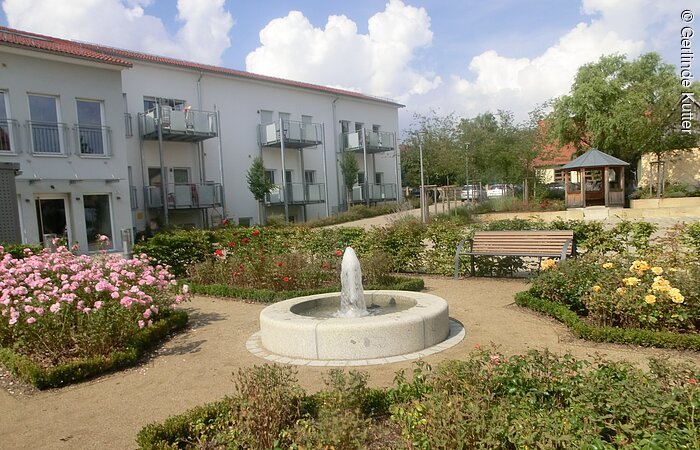 Ein kleiner Park mit Schotterwegen und bepflanzenten Beeten. In der Mitte ein kleiner Springbrunnen aus Stein. Im Hintergund ein langes Wohnhaus mit mehreren Parteien.