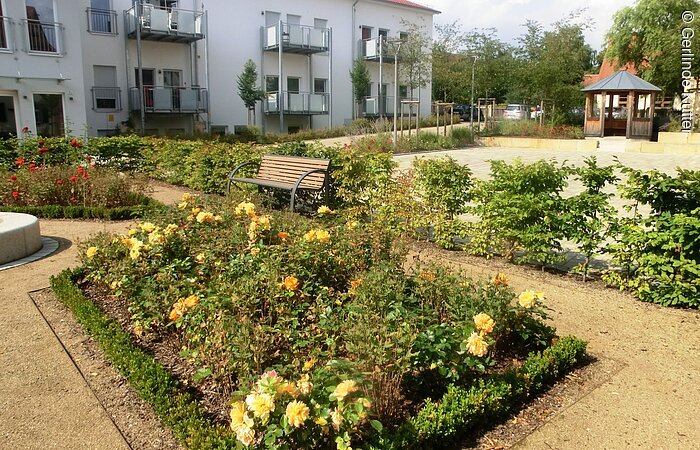 Viele Pflanzen blühen im Barockgarten. Grüne Flächen mit blühenden Blumen auf einem Schotterplatz. Im Hintergrund ein langes Wohnhaus mit einzelnen Wohungen. Jede Wohung hat einen Balkon. Ingesamt sechs Balkone.