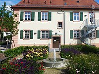 Ein Haus mit weißer heller gelber Fassade im barocken Baustil. An den Fenstern grüne fensterläden. Davor ein kleiner Park mit Springbrunnen, blühenden Büschen und einer Sitzbank. Umgeben ist der Garten von einer grünen Hecke.