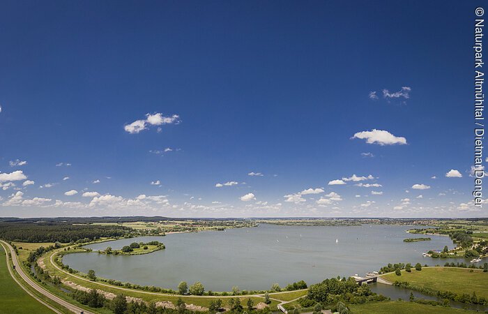 Panoramablick auf einen See mit Uferwegen, Wiesen und blauem Himmel mit vereinzelten Wolken.