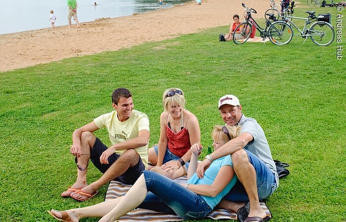 Zwei Frauen und zwei Männer sitzen auf einer Decke auf einer Wiese am Ufer des Altmühlsee und unterhalten sich. Im Hintergrund sind Fahrräder, weitere Menschen sowie ein Strand und der Altmühlsee zu erkennen.