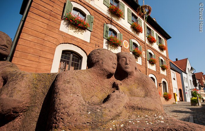 Rathaus Windsbach Blick und untern auf zwei Figuren eines Steinbrunnen. Die zwei Figuren halten Hände. Im Hintergund ein großes Haus mit Steinfassade. An den Fenstern grüne Fensterläden und Blumenkästen mit roten Blumen.