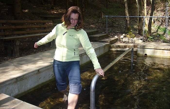 Langsam watet eine Bescuherin barfuß durch das eiskalte Wasser des Kneippbeckens in Heidenheim. Hinter dem Kneippbecken eine Holzbank. Dahinter Wald.