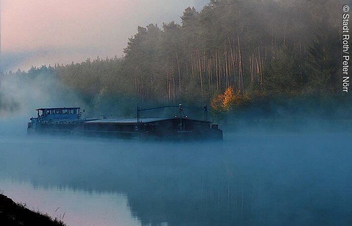 Ein Frachtschiff auf dem Main-Donau-Kanal. Das Wasser leuchtet hellblau. Über dem Kanal hängt Nebel. Am hinteren Ufer erstreckt sich ein Wald der im Nebel liegt.