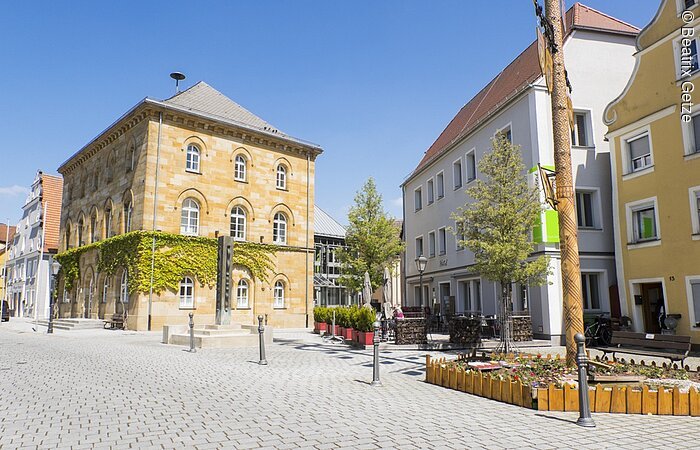 Rathaus Wassertrüdingen Eine Innenstadt mit großen Häusern. Im Fokus liegt ein Steinhaus mit drei Ebenen. Davor ein gepflasterter Platz. Rechts davon Wohnhäuser.