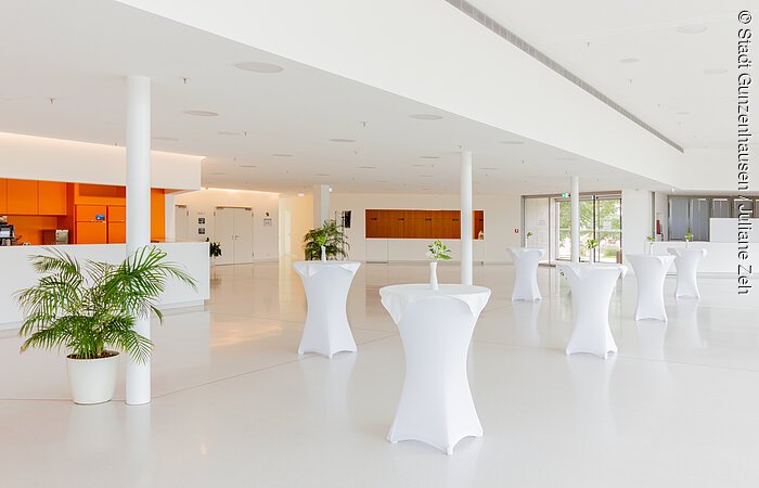 Das Foyer der Stadthalle Gunzenhausen Heller Raum mit Stehtischen, weiß bezogen, Pflanzen in Töpfen, oranger Bereich links, große Fenster rechts.
