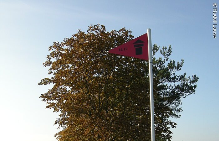 Eine rote Fahne weist den Weg des Panorama-Rundwanderwegs. Die Fahne steht auf einer grüne Wiese an einem Wanderweg. Dahinter ein Baum mit braunen Blättern.