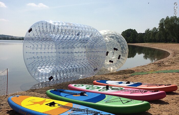Vier SUP-Boards liegen an einem Snadstrand. Dahinter treiben zwei doppelhüllige transparente Kugel im Wasser.