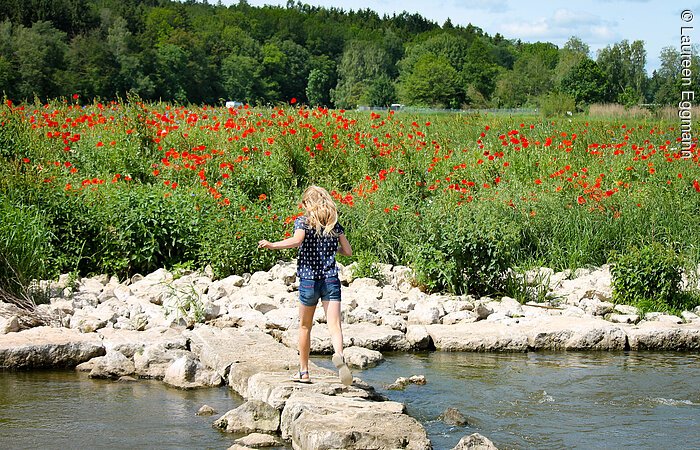 Rote Mohnfelder Ein junges Mädchen läuft über eine Steinreihe die durch einen Fluss führt. Dahinter ein rot blühendes Mohnblumenfeld.