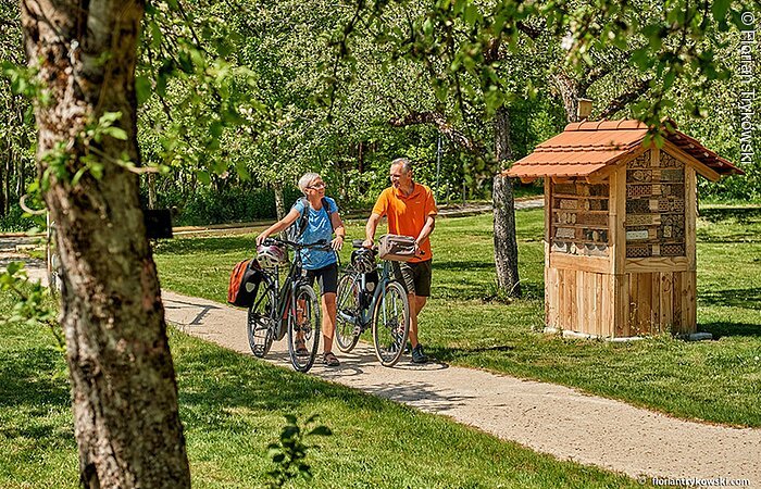 Spaziergang mit dem Fahrrad Zwei Radler schieben ihre Fahrräder durch einen kleinen Park entlang eines Schotterwegs. Links und Rechts Wiesen und Bäume sowie ein Insektenhotel aus Holz.