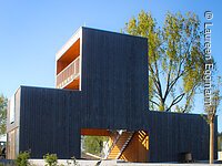 Aussichtspavillion Ein offenes Gebäude mit Holzfassade in Form. Der Pavilion besteht aus drei Blöcken. Davor eine grüne Wiese.