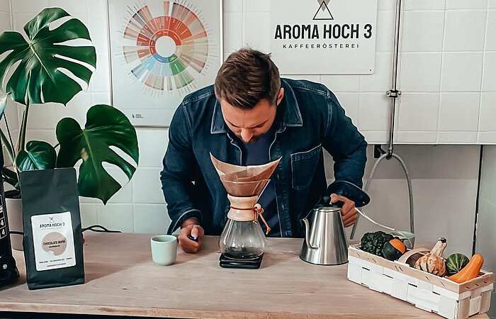 Kaffeegenuss erleben bei Aroma Hoch 3 Ein junger Mann steht an einer Theke. Er beugt sich über eine KAffeefilter und riecht daran. In der linken Hand eine Kanne, neben der rechten Hand steht eine Tasse. An der Wand dahinter hängt ein weißes Schild mit schwarzer Schrift: Aroma Hoch 3