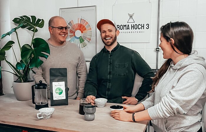 Großartiger Kaffee für großartige Leute bei Aroma Hoch 3 Zwei Männer und eine Frau stehen um eine Theke und unterhalten sich. Auf der Theke eine Packung Kaffee und drei Kaffeetassen. An der Wand hinter der Gruppe hänt ein Schild auf welchem im schwarzer Schrift steht: Aroma Hoch 3
