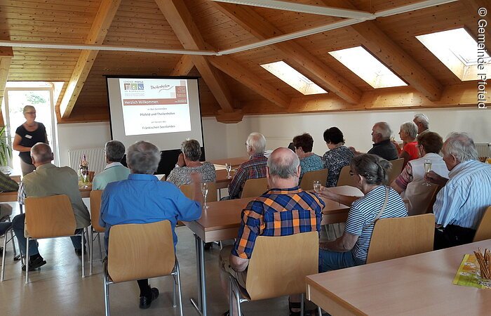 Eine große Gruppe Menschen sitzt auf Holzstühlen und blickt auf eine Leinwand mit Präsentation. Links neben der Leinwand steht eine Frau und sprciht. Die Decke des Raumes ist mit Holz verkleidet.