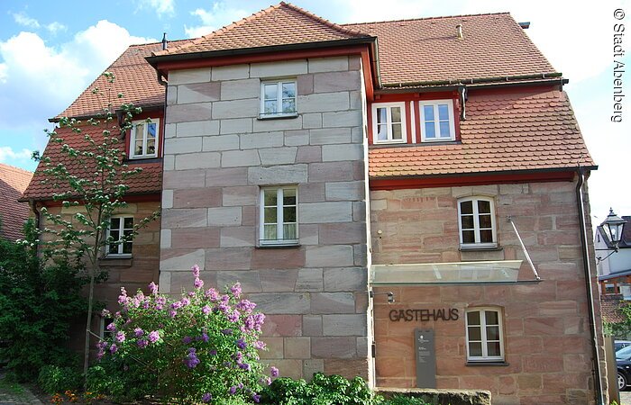Ein großes Haus aus Stein mit rotem Ziegeldach. Direkt vor dem Haus grüne Büsche und Blumen.