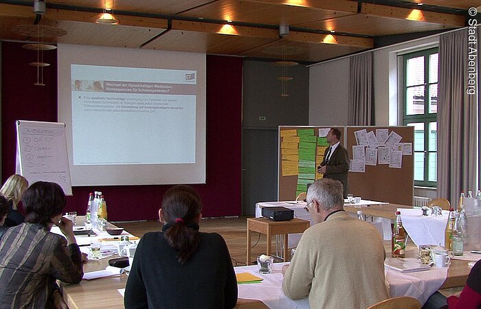 Eine Tagung im Abenberger Hotel "Burg Abenberg". Ein Mann steht neben einer weißen Leinwand mit Präsenation. Vor ihm sind Tische in U-Form angeordnet. An den Tischen sitzen die Zuhörer und lauschen dem Vortrag.