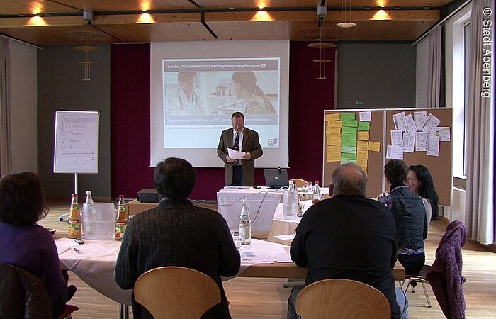 Eine Tagung im Abenberger Hotel "Burg Abenberg". Ein Referent steht neben einer weißen Leinwand mit Präsenation. Vor ihm sind Tische in U-Form angeordnet. An den Tischen sitzen die Zuhörer und lauschen dem Vortrag.