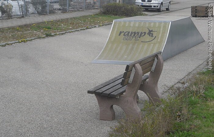 Skateanlage mit einer Skateboardrampe. Im rechten Bildauschnitt steht vor der Rampe eine Sitzbank. Im Hintergund ist ein Zaun und zwei parkende Autos zu erkennen.