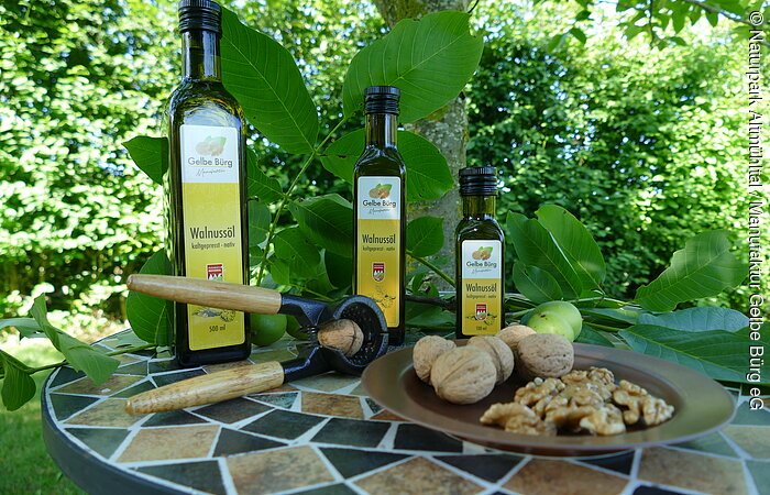 Auf einem runden Mosaiktisch stehen drei Flaschen Walnussöl in verschiedenen Größen mit einem Teller Walnüsse und Walnusskerne. Der Tisch steht vor einem grünen Gebüsch.