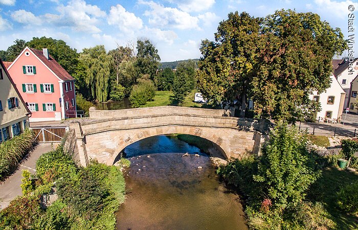 Luftaufnahme einer Steinbrücke. Die Brücke fällt über einen Fluss. Links und rechts Wohnhäuser.