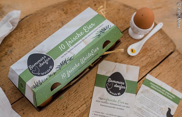 Ein geschlossener Eierkarton steht auf einem Holzbrett. Rechts davon ein Eierbecher mit Ei und Löffel. Auf der Eierschachtel steht Bammersdorfer Weide Ei.