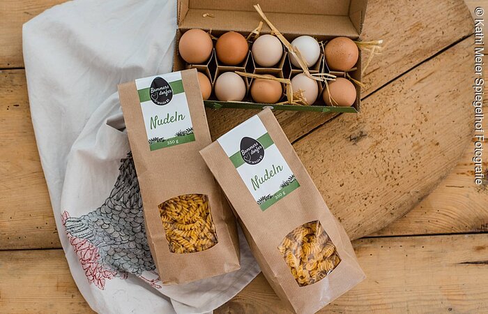 Produktpräsentation von Nudeln und Eiern platziert auf Holz. Ein offener, gefüllter Eierkarton. Davor liegen zwei Packungen Nudeln.