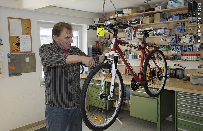 Ein Mann repariert ein rotes Fahrrad, das von der Decke hängt. Er steht in einer Fahrradwerkstatt
