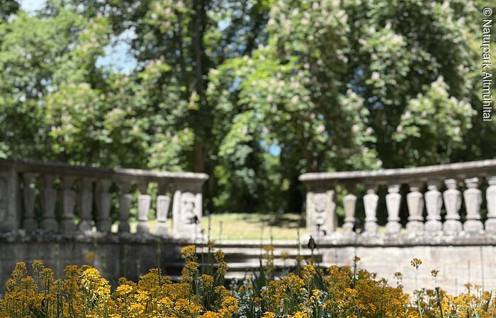 Ein Beet mit Planzen im Vordergrund wird von einer Mauer abgegrenzt. Ein steinerner Weg führt auf eine höhere Ebene mit Bäumen