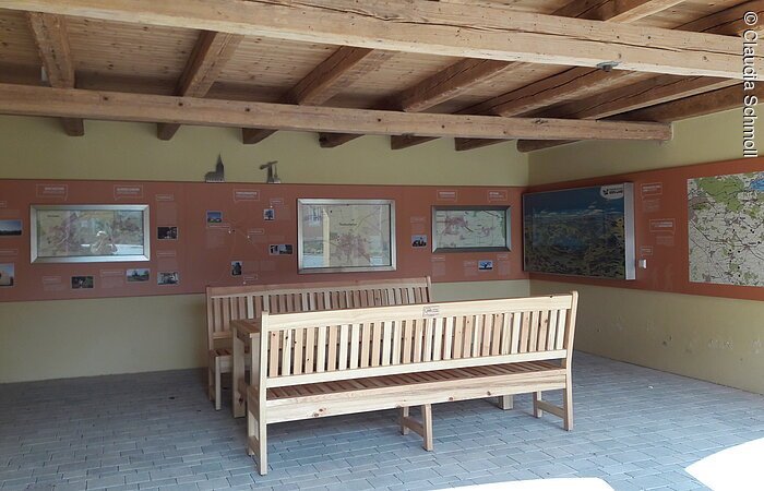 Innenraum eines offenen Häuschen. Darin stehen eine Sitzgruppe aus Holz. An der gelb-roten Wand hängen Informationen zum Limes.