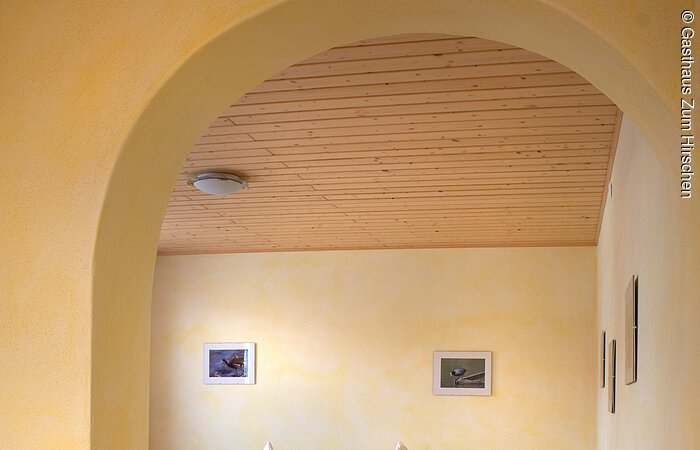 Durch einen Torbogen in der Wand fällt der Blick auf ein Doppelbett aus Holz mit blumiger Bettwäsche. Ein hellgelber Raum mit Dekce und Boden aus Holz.