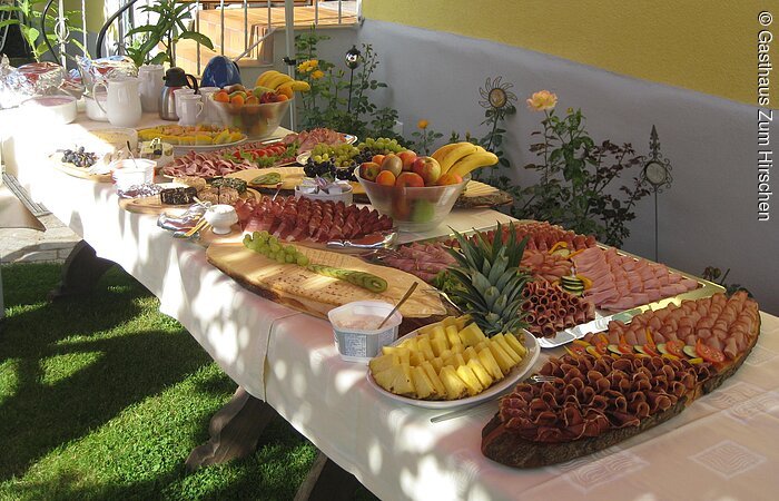 Großes Buffet mit verschiednen Leckereien. Ein langer Tisch mit weißer Tischdecke steht in einem Garten an einer gelben Hauswand. Auf dem Tisch viele vershciedne Platten mit Obst, Wurst oder Käse.