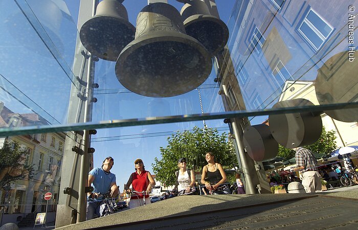 Blick auf die Glocken des verglasten Glockenturms in Gunzenhausen. Dahinter stehen vier Radfahrer und betrachten die Glocken.