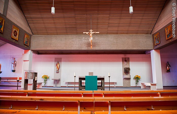Der Innenraum einer katholischen Kirche. Bänke aus Holz. Dahinter eine Altar. Darüber hängt ein Kreus mit Jesus Christus. Die Decke ist holzverkleidet.