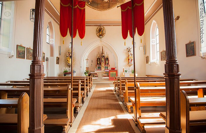 Der Innenraum einer katholischen Kirche. Sitzbänke aus Holz stehen in zwei Reihen entlang des Kirchenschiffs. Am Ende ein Taufbecken. Dahint ein prunkvoller Altar mit Figuren.
