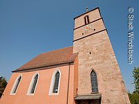 Außenansicht einer katholischen Kirche mit Kirchenschiff und Kirchturm. Das Kirchenschiff hat eine oragene Fassade und hohe Fenster. Der Kirchturm ist aus Stein gemauert.