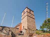 Blick von unten auf den Steinturm einer katholischen Kirche. Davor verläuft eine Mauer aus Stein.