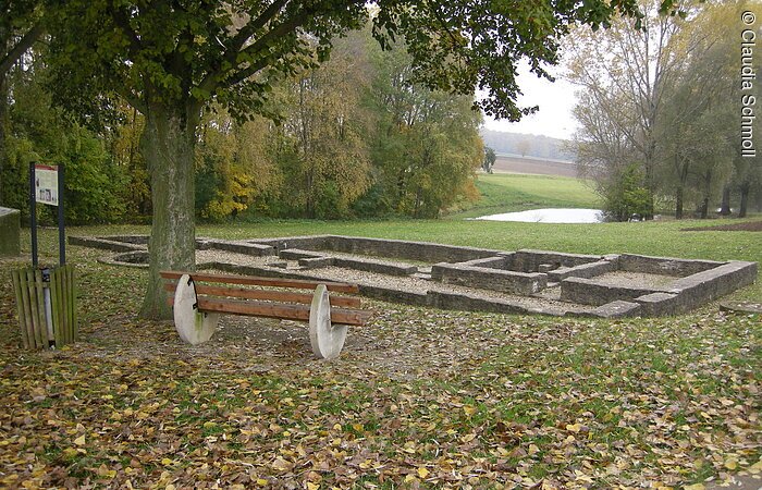 Eine Sitzbank unter einem Baum auf einer Wiese mit gelbem Laub. Von der Bank aus hat man Blick auf den den Umriss eines alten Römerbads. Im Hintergund Bäume und Sträucher.
