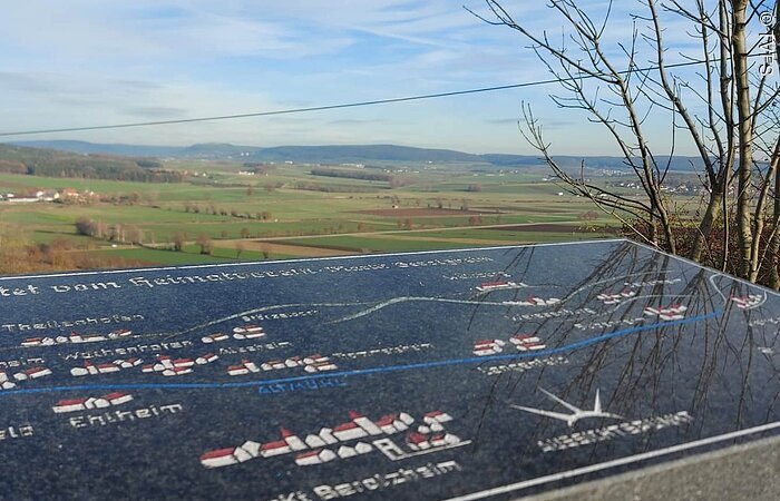 Im Vordergrund eine Steintafel. Dahinter eine weite grüne Landschaft. Felder und Bäume.