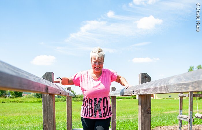 Motorikpark Eine Frau mit pinkem Shirt und schwarzer Hose balanciert über ein in der Luft gespanntes Seil. Dabei hält sie sich links und rechts an einem Holzgeländer fest.