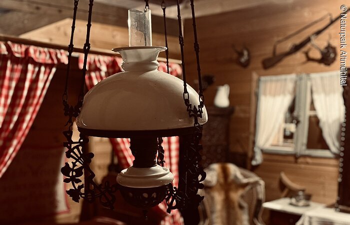 In der Mitte des Bildes hängt eine Öllampe von der Decke über einem Tisch. Im Hintergrund ist eine Fensterbank mit roten Vorhängen zu sehen.