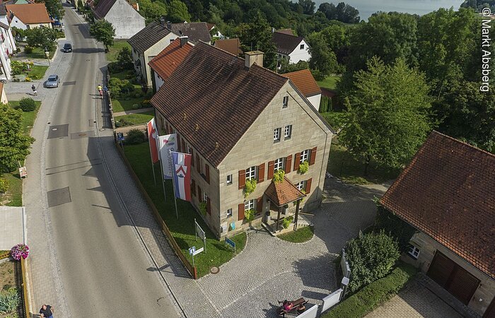 Ein Luftbild eine Hauses aus Stein mit roten Fensterläden. An der Hauswand hängen drei bunte Fahnen. Das Haus steht an einer Straße in einem Wohnegbiet.