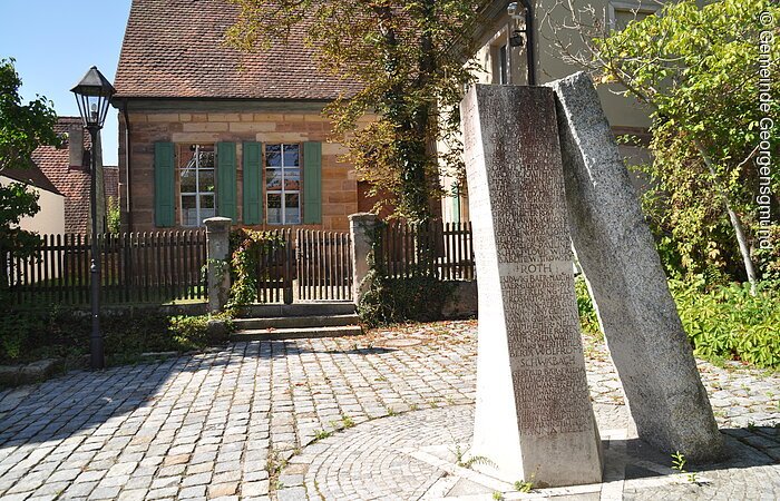 Ein grauer Gedenkstein auf einer gepflasterten Fläche. Dahinter ein Steinhaus mit grünen Fensterläden.