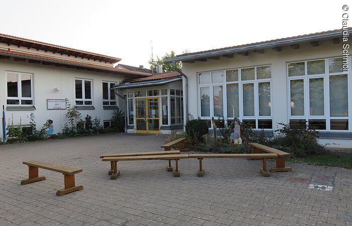 Außenansicht eines ebenen Hauses mit hohen Fnstern. Weiße Fassade und rotes Ziegeldach. Davor ein gepflasteter Hof mit fünf Sitzbänken aus Holz.