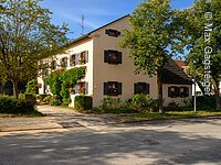 Das Familienhaus ist einen hellen Ton angestrichen und hat grüne Fensterläden, an den Fenstern befinden sich Blumenkästen mit Geranien. Um das Haus befinden sich verschiedene Sträucher und an der westlichen Häuserfront befinden sich Pflanzen