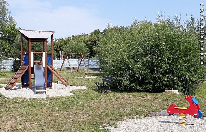 Eine blaue Rutsche mit Klettergerüst. Rechts davon ein großer grüner Busch. Davor ein Holzmotorrad zum wippen.