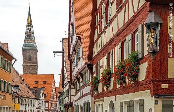 Lammswirtshaus in Wolframs-Eschenbach Blick in eine Altstadt. Rechts und links Häuserreihen von Fachwerkhäusern. Im Hintergund ragt ein Kirchturm mit bunten Ziegeln über den Dächern heraus.