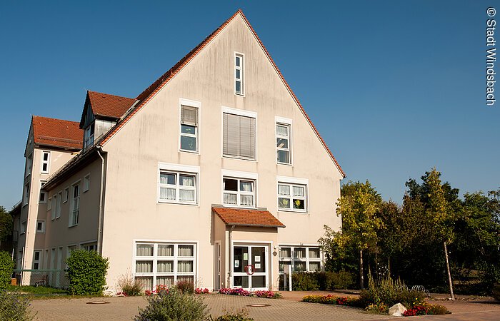 Stadtbücherei Windsbach Ein großes Haus mit vier Ebenen. Helle rosane Fassade mit weißen Fenstern. Rechts davon Bäume.