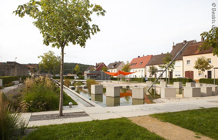 Sonnenuhrenpark Ein Park mit einem Teich. Im Teich sind kreisförmig Steinblöcke angeordnet. Sie bidlen eine Sonnenuhr. Um den Teich Sträucher und Blumen. Im Hintergund Wohnhäuser.